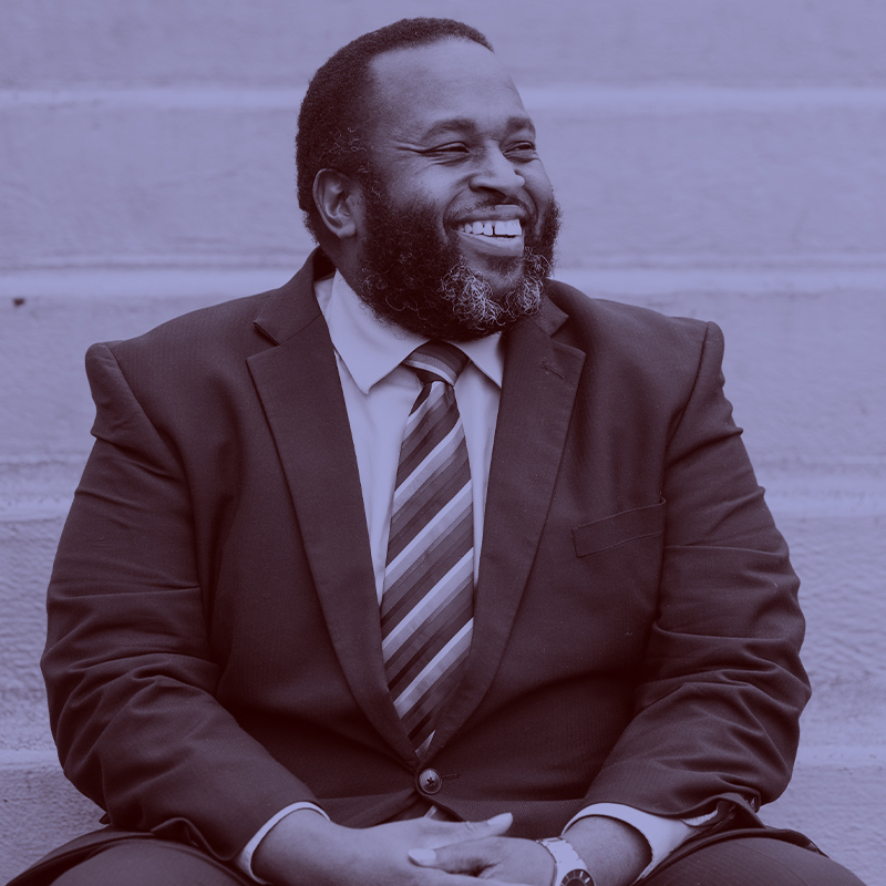 Photo: Eron Hackshaw, a Black man with a greying black beard wearing a navy blue suit, white collared shirt and striped tie, smiles and looks to the left as he sits on a set of cement stairs and looks to the right. A light purple filter overlay is placed over the photo.