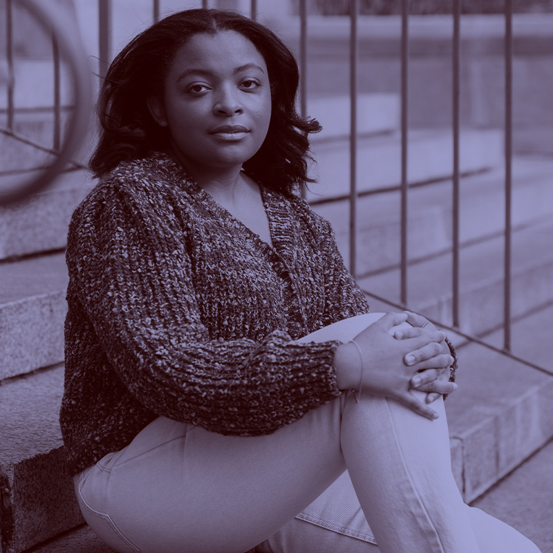 Photo: A Black woman with wavy, roller set hair, poses on a stoop with hands holding one knee up. A light purple filter overlay is placed over the photo.