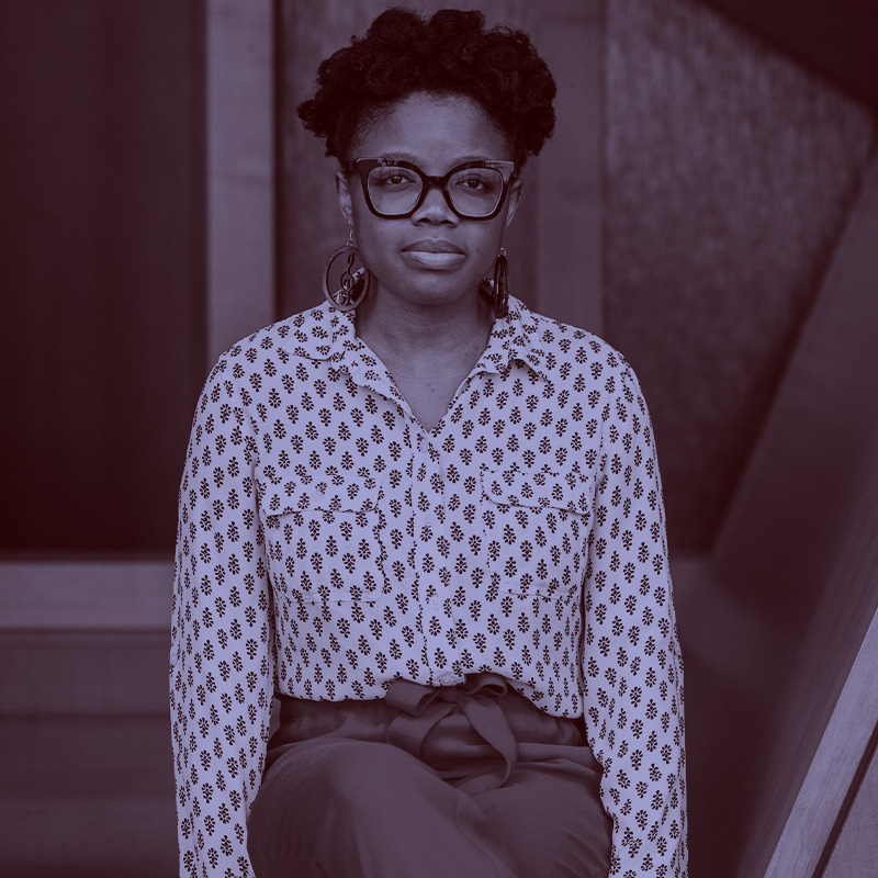 Photo: Elaine Nsoesie, a Black woman with a short afro hairstyle, wears glasses, a patterned collared shirt, and slacks. She sits with hands at sides as she poses for the photo. A light purple filter overlay is placed over the photo.