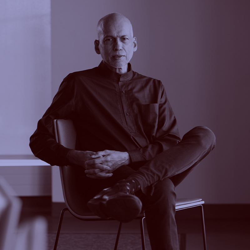 Photo: A black man with a  somber expression sits with hands in lap and one leg crossed over the other. He wears a black long-sleeved collared shirt and black framed glasses. A light purple filter overlay is placed over the photo.