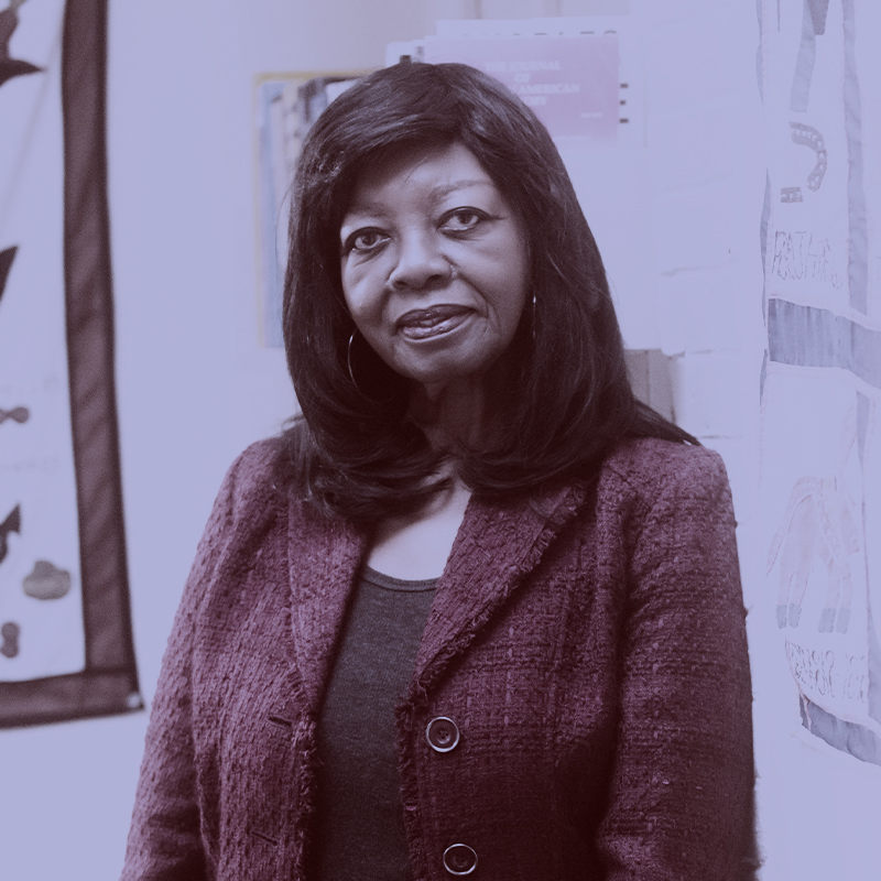 Photo: Joyce Hope Scott, a Black woman with shoulder-length straight black hair and wearing a black top and red tweed blazer, poses for a photo. A light purple filter overlay is placed over the photo.