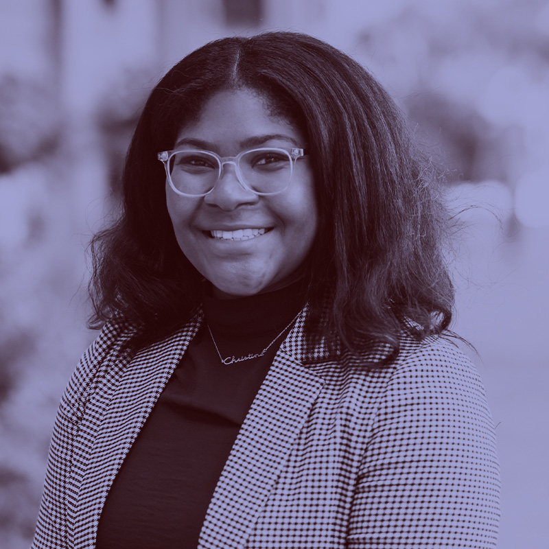 Photo of Christine Slaughter. A young Black woman wearing glasses, a burgundy sweater, and twead jacket, smiles for the camera. A light purple filter overlay is placed over the photo.