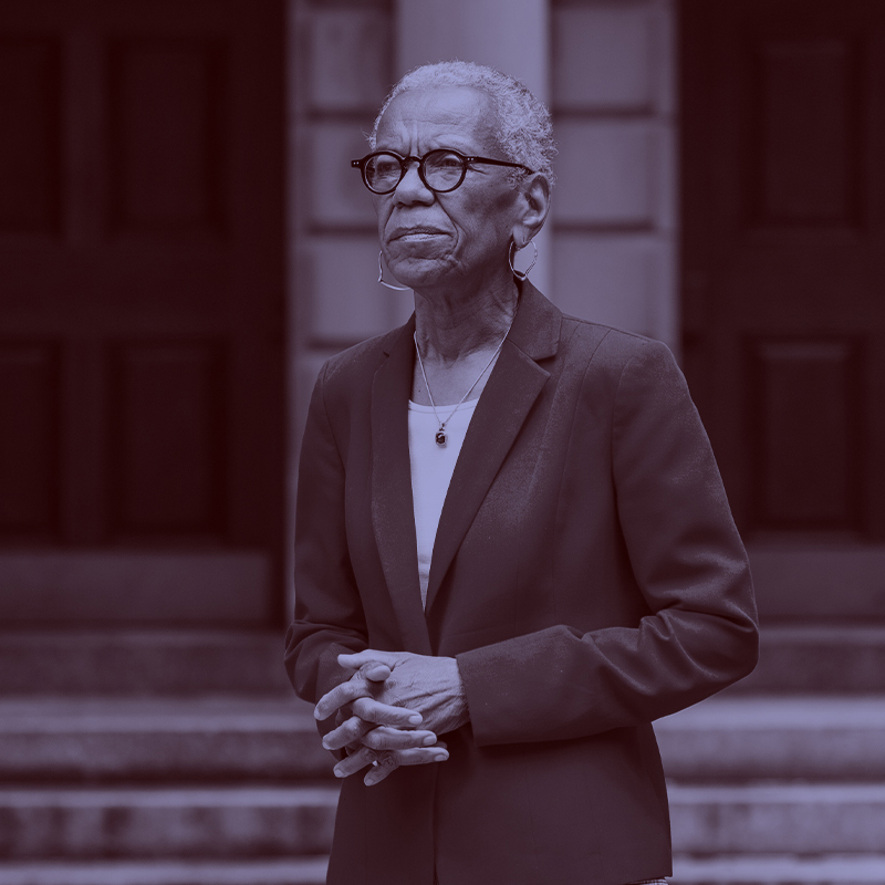 Photo: Andrea Taylor, a Black woman with short silver-white hair, looks off to the left with hands clasped in front of her. She wears glasses, a red blazer and white blouse. A light purple filter overlay is placed over the photo.