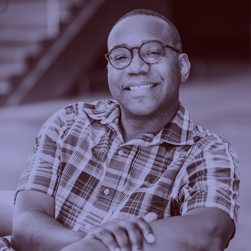 Photo: Vincent Stephens, a smiling Black man wearing glasses and a plaid shirt, poses for a photo on an outdoor bench. A light purple filter overlay is placed over the photo.