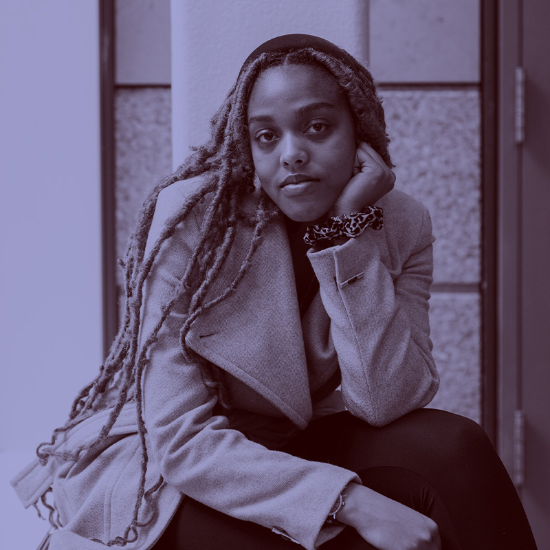 Photo: Deborah Williams, a young Black woman with long dreads and wearing a tan blazer poses with head in hand. A light purple filter overlay is placed over the photo.