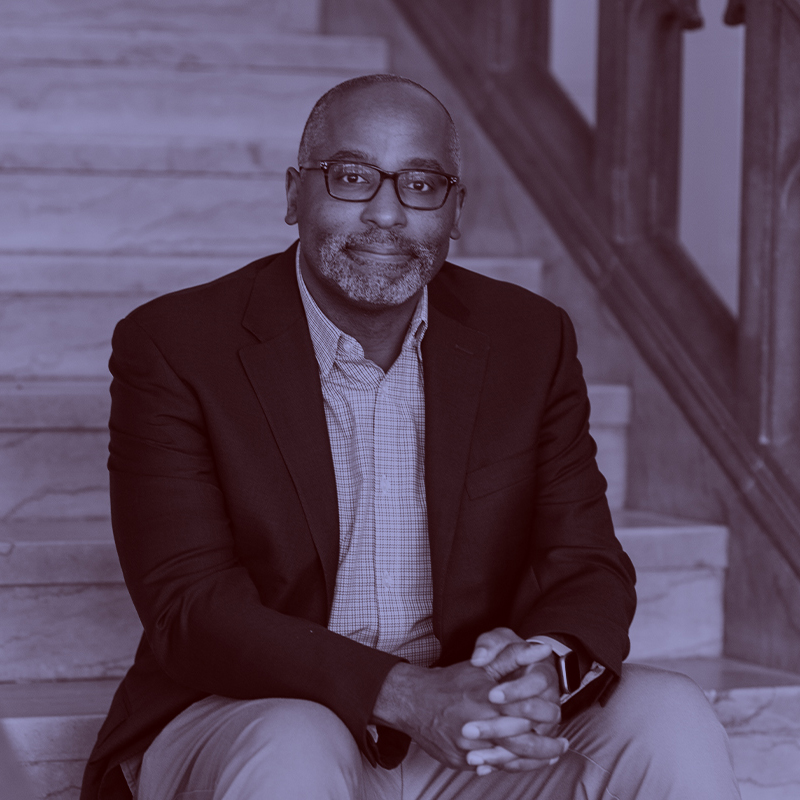 Photo: CFA Dean Harvey Young. A black man wearing glasses, a collared shirt, black blazer, and slacks, poses and sits on a set of stairs. A light purple filter overlay is placed over the photo.