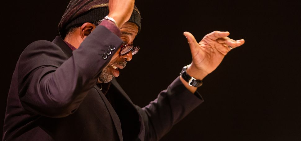 Photo: Bill Banfield (STH'88) conducts a concert with his Imagine Orchestra at the Edward M. Pickman Concert Hall Longy School of Music, Friday, November 11, 2022. A Black mean wearing glasses, a purple and black knit cap, and black blazer, has his hands raised in the air as he conducts.