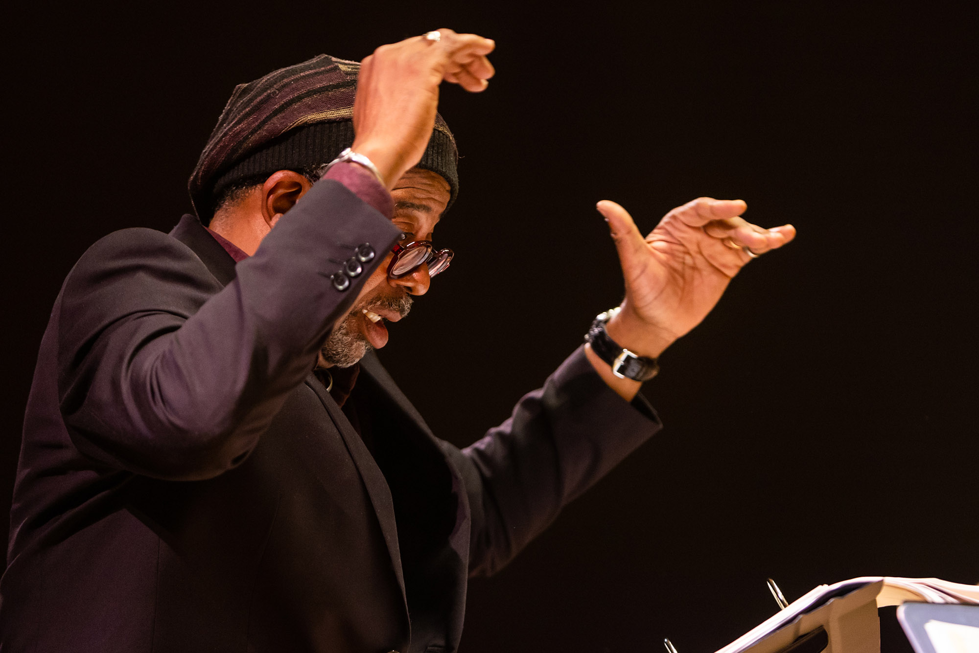 Photo: Bill Banfield (STH'88) conducts a concert with his Imagine Orchestra at the Edward M. Pickman Concert Hall Longy School of Music, Friday, November 11, 2022. A Black mean wearing glasses, a purple and black knit cap, and black blazer, has his hands raised in the air as he conducts.