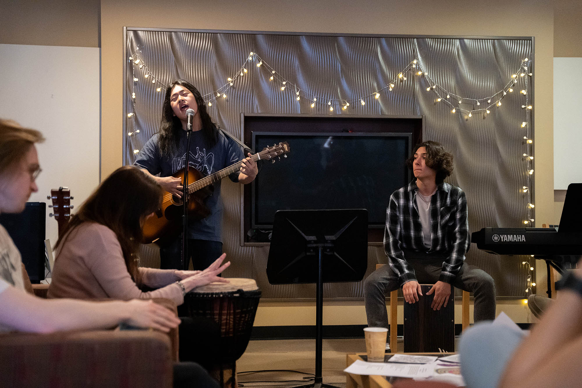 Photo: Andrew Lee (CGS'26) (left) and Eli Gately (CGS'26) (right) perform at the CGS Art Show and Coffee House. A young man with long black hair sings passionately into a mic as he plays a guitar. A student to his right sits and plays a large hand drum. Another student in the audience plays along on another drum.