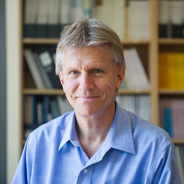 Photo: Headshot of Davidson Hamer. An older white man with brown gray hair and wearing a light blue collared shirt smiles and poses in front of a stack of books.