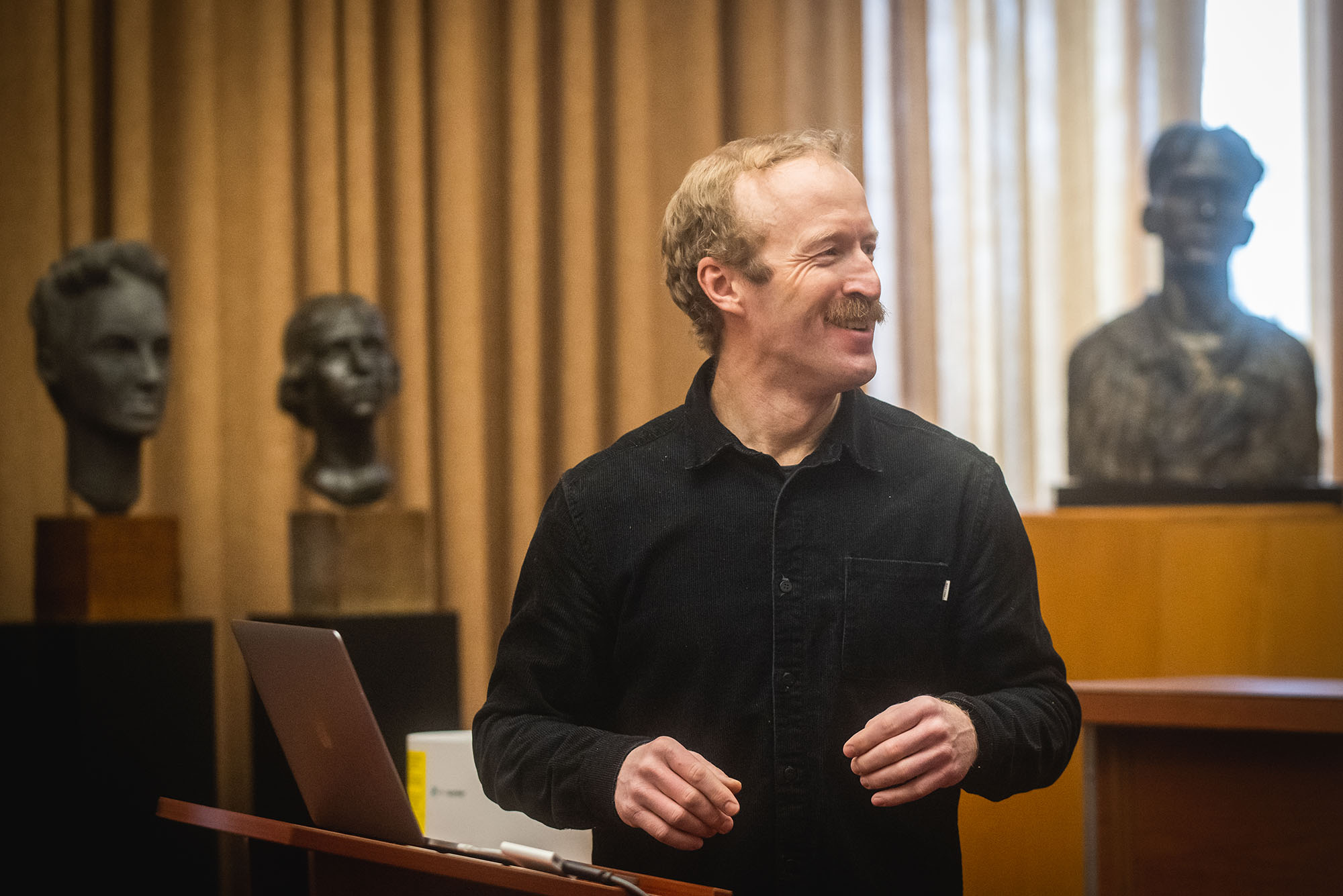 Photo: Griffin Post stands at a podium and looks to the right. A white man wearing a black long-sleeved collared shirt stands in front of a podium where a silver laptop rests as he smiles and looks to the right.