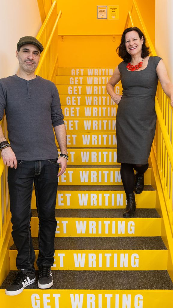 Photo: Artistic director Dariel Suarez (GRS’12) (left) and founder and executive director Eve Bridburg (GRS’97) stand and pose on a set of bright yellow stairs. Each step has the words "Get writing" printed on them in white. A latino man wearing a grey beret, grey shirt, and black pants stands with one arm resting on the railing to his left and smiles. To his right, a white woman wearing a grey dress, black pantyhose, and red chunky necklace smiles and stands with one arm resting and poised on the railing to her right.