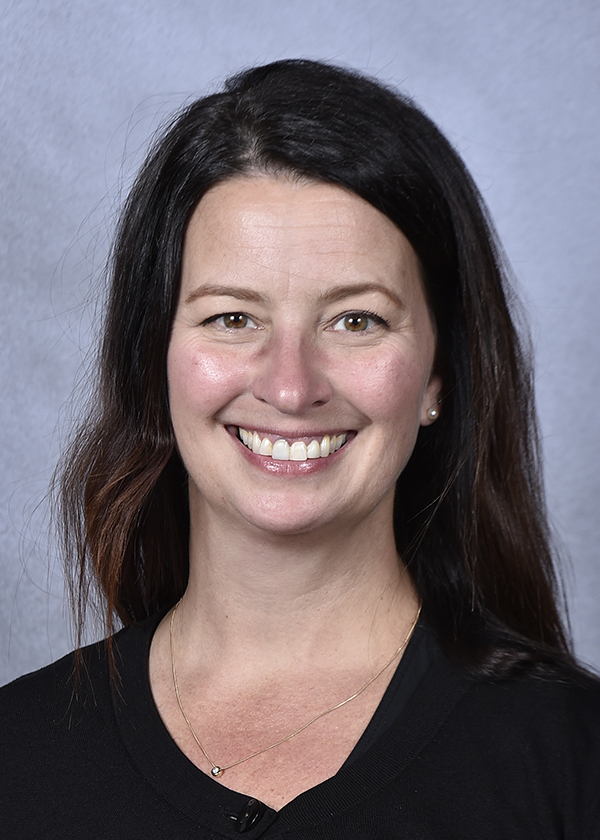 Photo: Headshot of Tracy Marek, a white woman with long dark brown hair and wearing a black top smiles and poses in front of a light blue backdrop.
