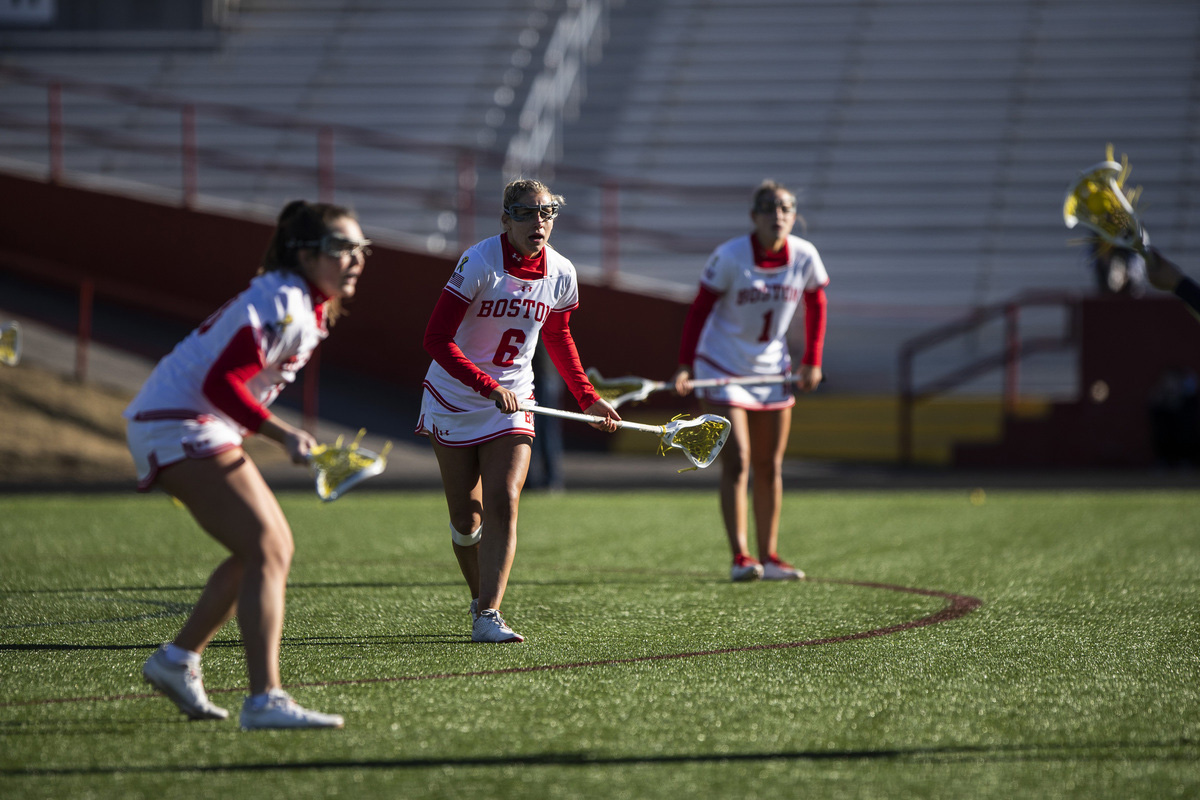Photo: BU cocaptain Maddie MacMaster (CGS’20, Questrom’22, Questrom’26) scouts out a Merrimack attack during the team’s season opener February 11. Three young women wearing white and scarlet red BU women's lacrosse uniforms crouch with sticks in front of them as they get in ready stances on the lacrosse field.