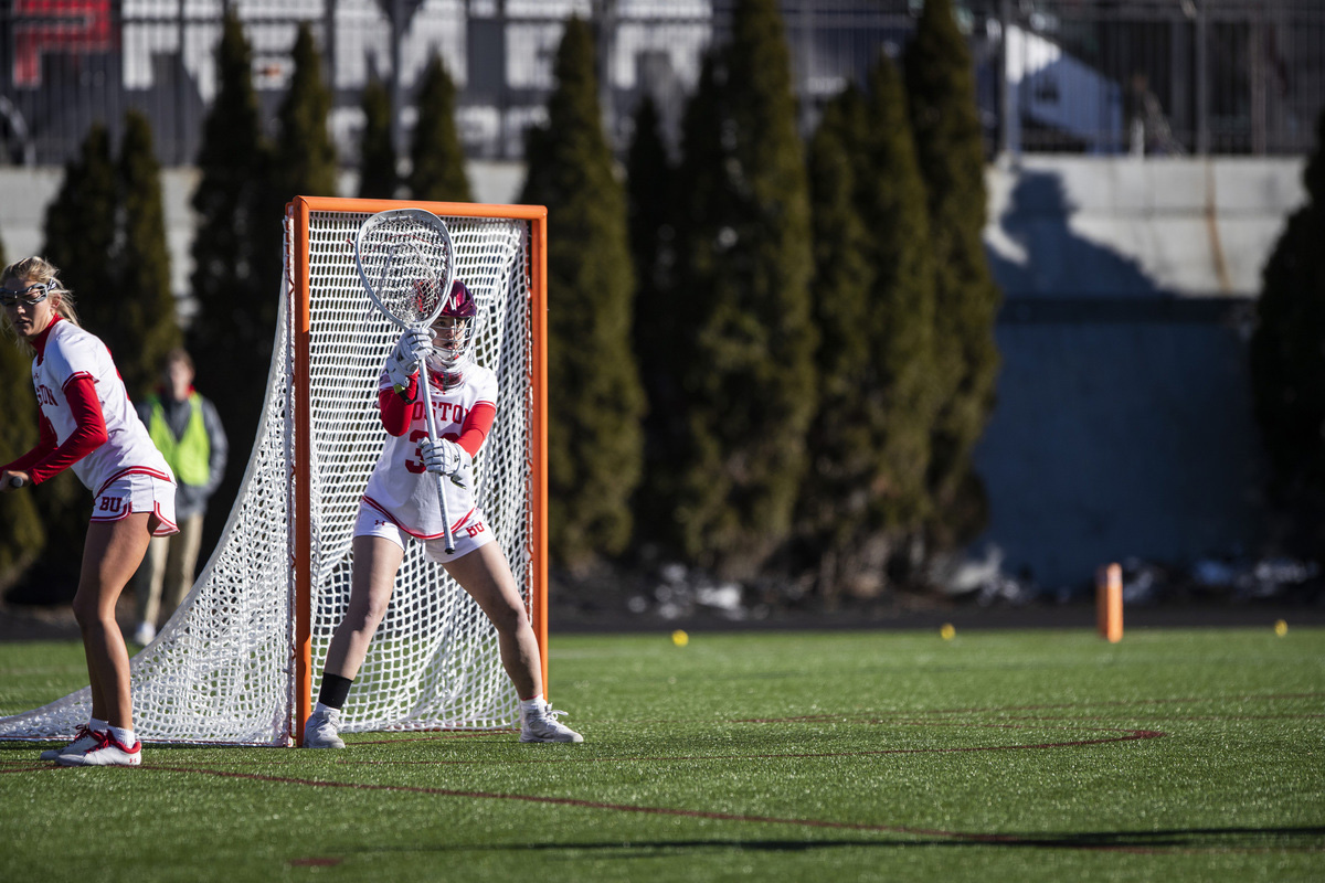Photo: BU cocaptain and goaltender Reilly Agres (CAS’24, Sargent’24) protects the cage against Merrimack on February 11. A young white woman wearing a white and scarlet red Bu women's lacrosse uniform holds up her lacrosse stick in front of the goal.