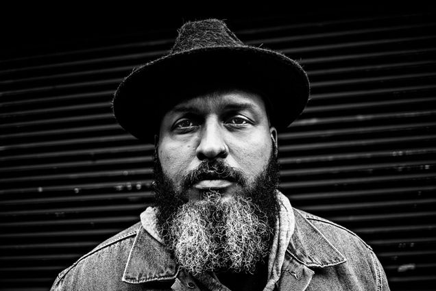 Photo: A black and white photo of a Black man with a long, peppered beard. He has a nose piercing in his left nostril. He was a collard shirt, a sweater overtop, and a flat-brimmed hat.