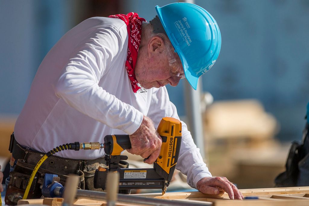 Photo: President Carter, at 93 years of age, drills into wood for habitat for humanity. He wears a blue hard-hat, red-bandana, and a white long-sleeve shirt.