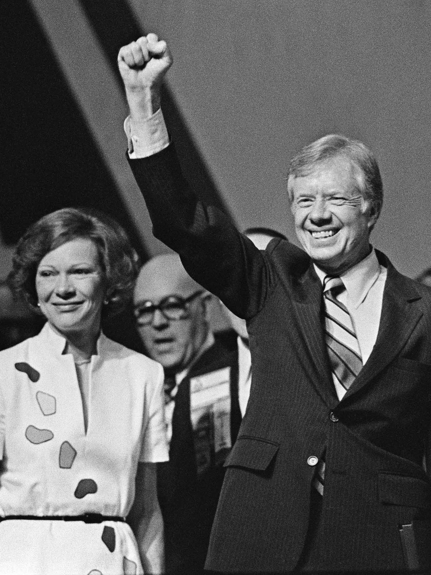 Photo: President Carter and Rosalynn Carter stand after President Carter gave a speech. He has his hand raised in a fist. They are smiling, him with his teeth and her with her mouth. It's a black and white photo.