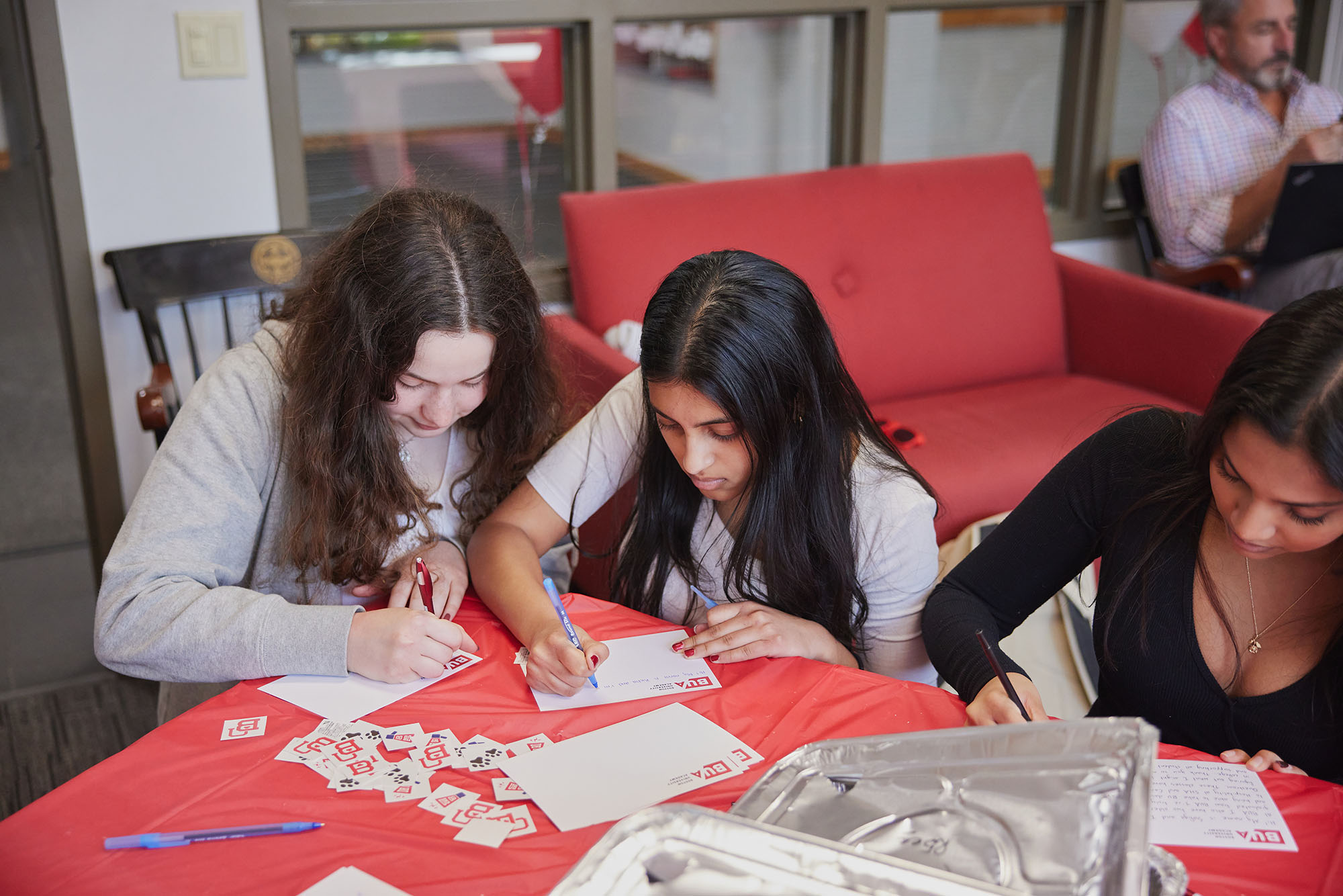 Photo: Three young students writing on white notecards on top of a table covered with a red tablecloth.
