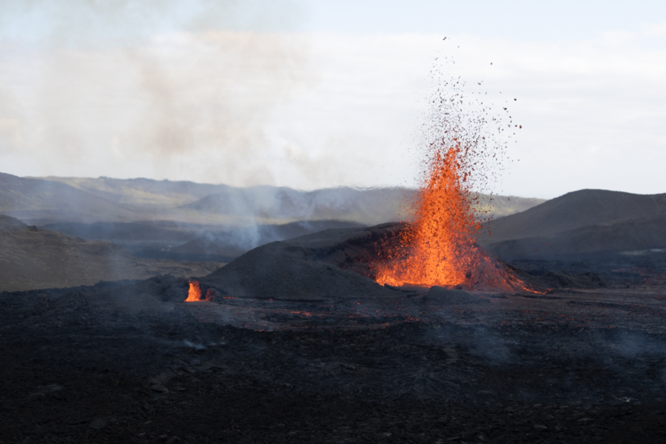 Photo: Fagradalsfjall volcano erupts as red lava escapes from new rifts in an adjacent valley. A black, rocky volcano with a large burst of lava shooting into the air is shown.