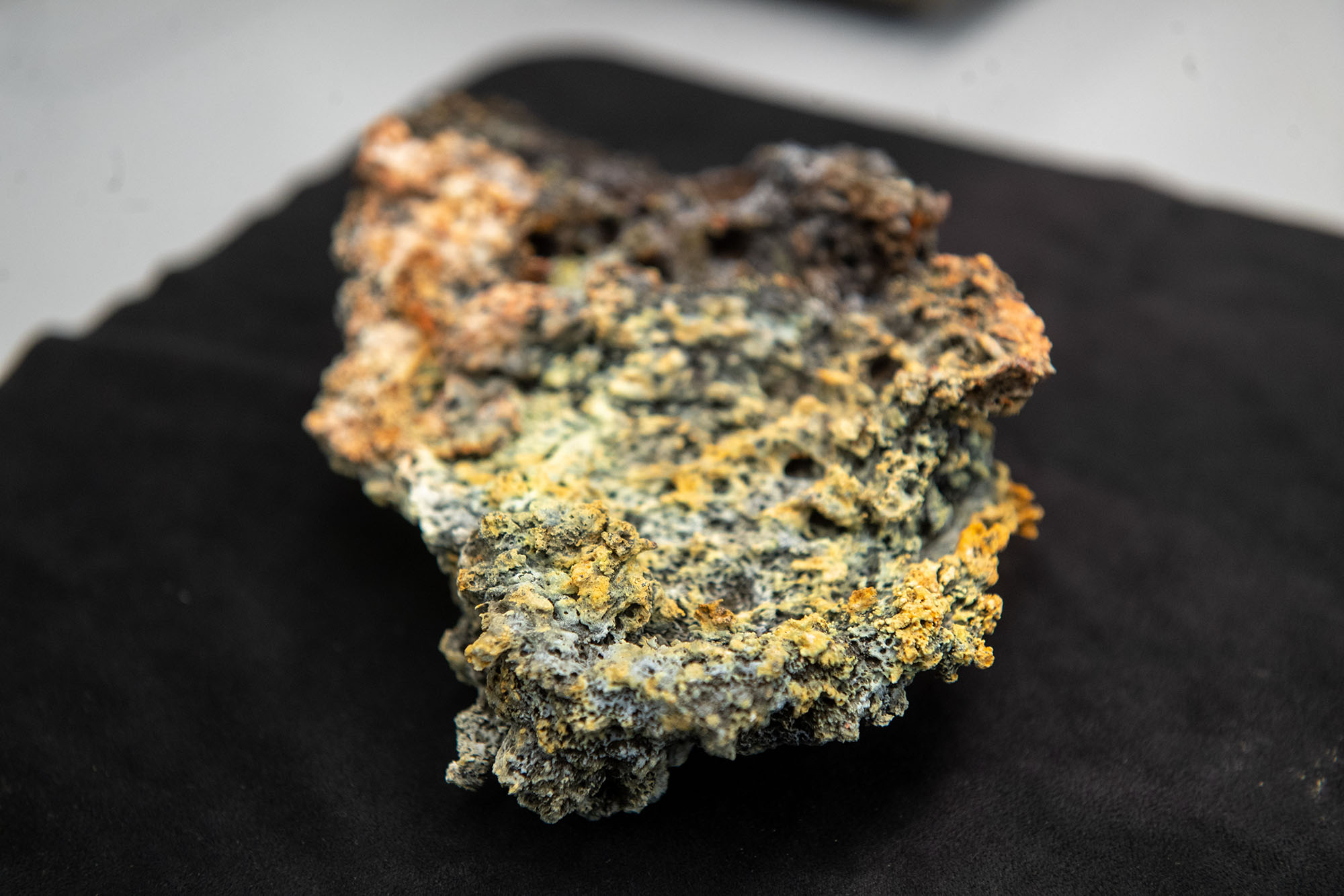 Photo: Closeup of a yellow, orange, grey, and black colored volcanic rock is shown sitting on a black cloth.
