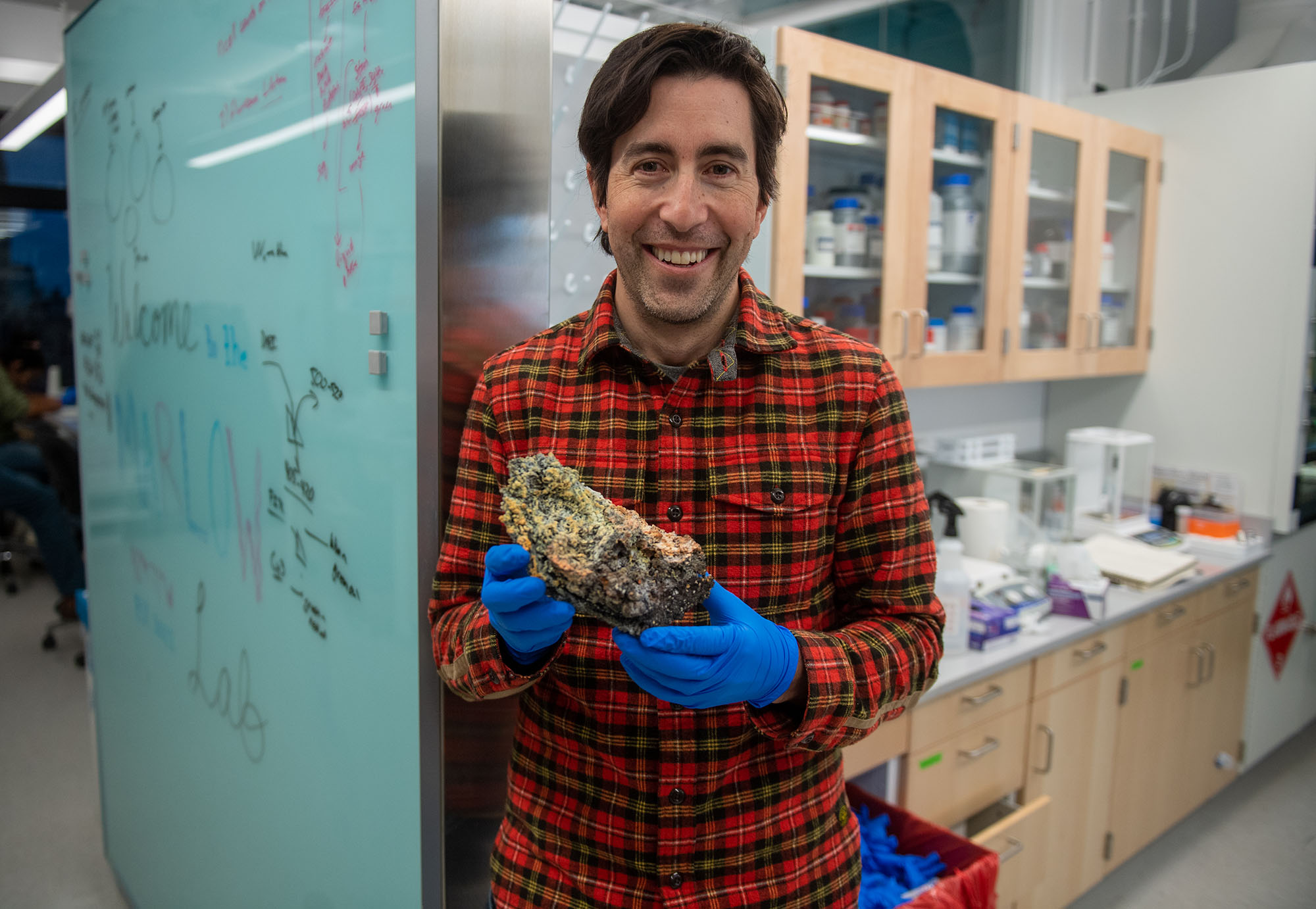 Photo: Jeffrey Marlow, a white man with dark brown hair and wearing a red, black and yellow plaid shirt and blue medical gloves holds up a volcanic rock as he poses in his lab.