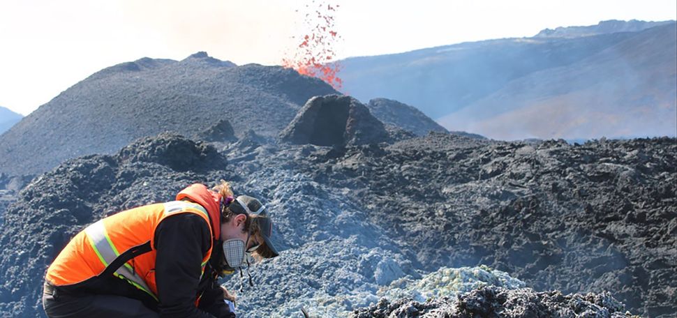 Photo: Iceland’s Fagradalsfjall volcano erupts in the background as Peter Schroedl, a person wearing black clothes, a face mask, orange neon vest collects specimens from volcanic rocks in the foreground.