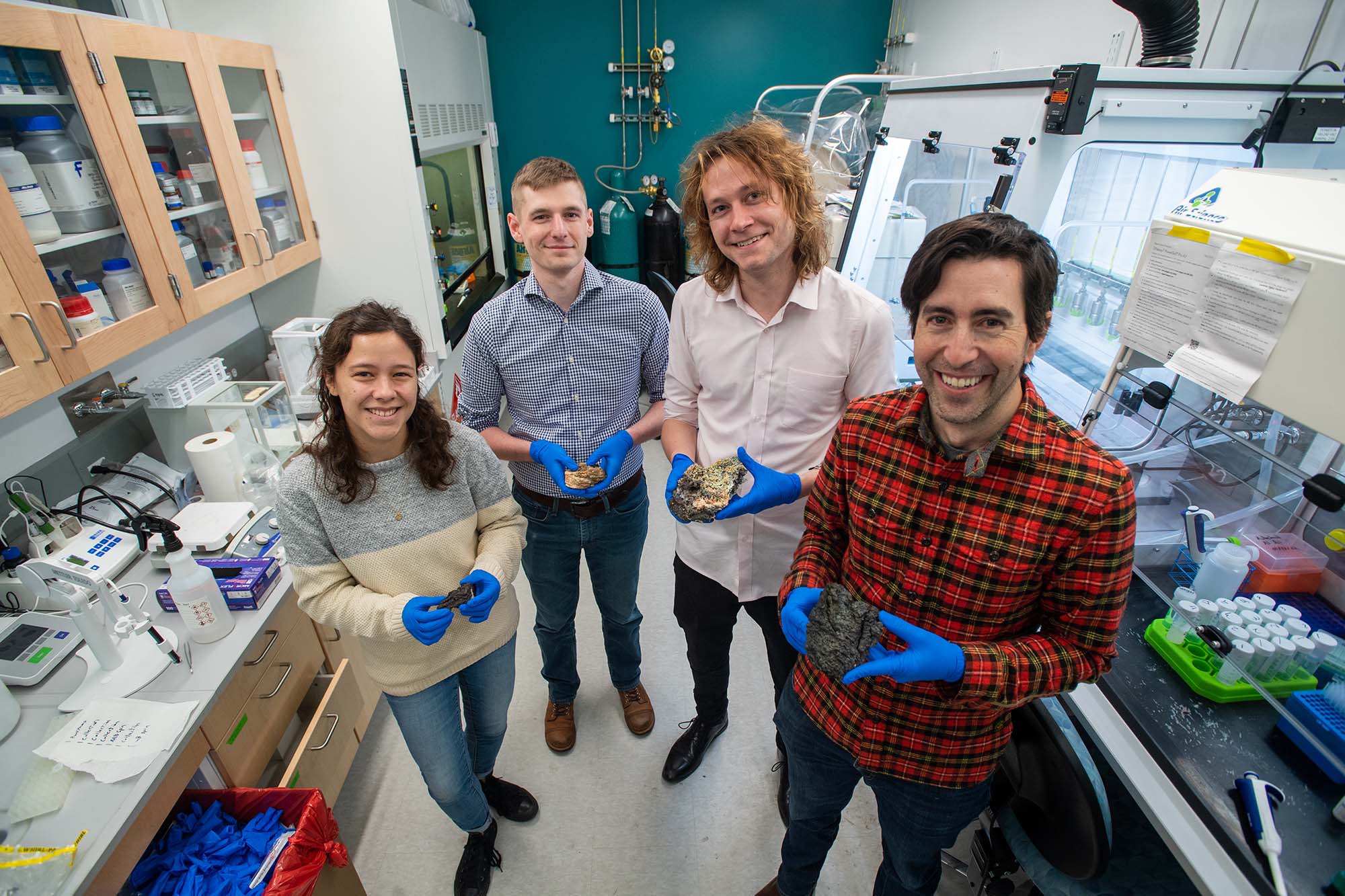 Photo: (from left) Erin Frates, Dylan Mankel, Peter Schroedl, and Jeffrey Marlow in the lab holding pieces of igneous rock collected from Fagradalsfjall, Iceland. A group of four white people wearing blue medical gloves hold pieces of volcanic rock as they stand and pose in their lab.