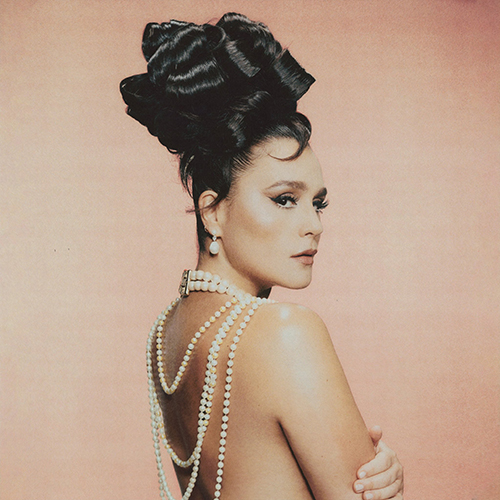 Album cover of Jessie Ware's "That! Feels Good!". Art shows Jessie Ware posing in front of a peachy background. Her hair is styled high in an elaborate bun. She's topless with her back turned to the camera as a large strong of pearls hangs from her neck and back in multiple layers.