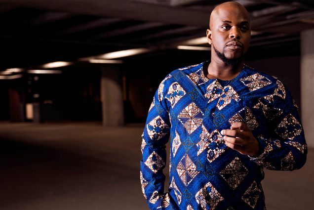 Photo: Patrick Dailey, a bald black man wearing an ornately designed sweater, poses for a portrait in a parking garage.