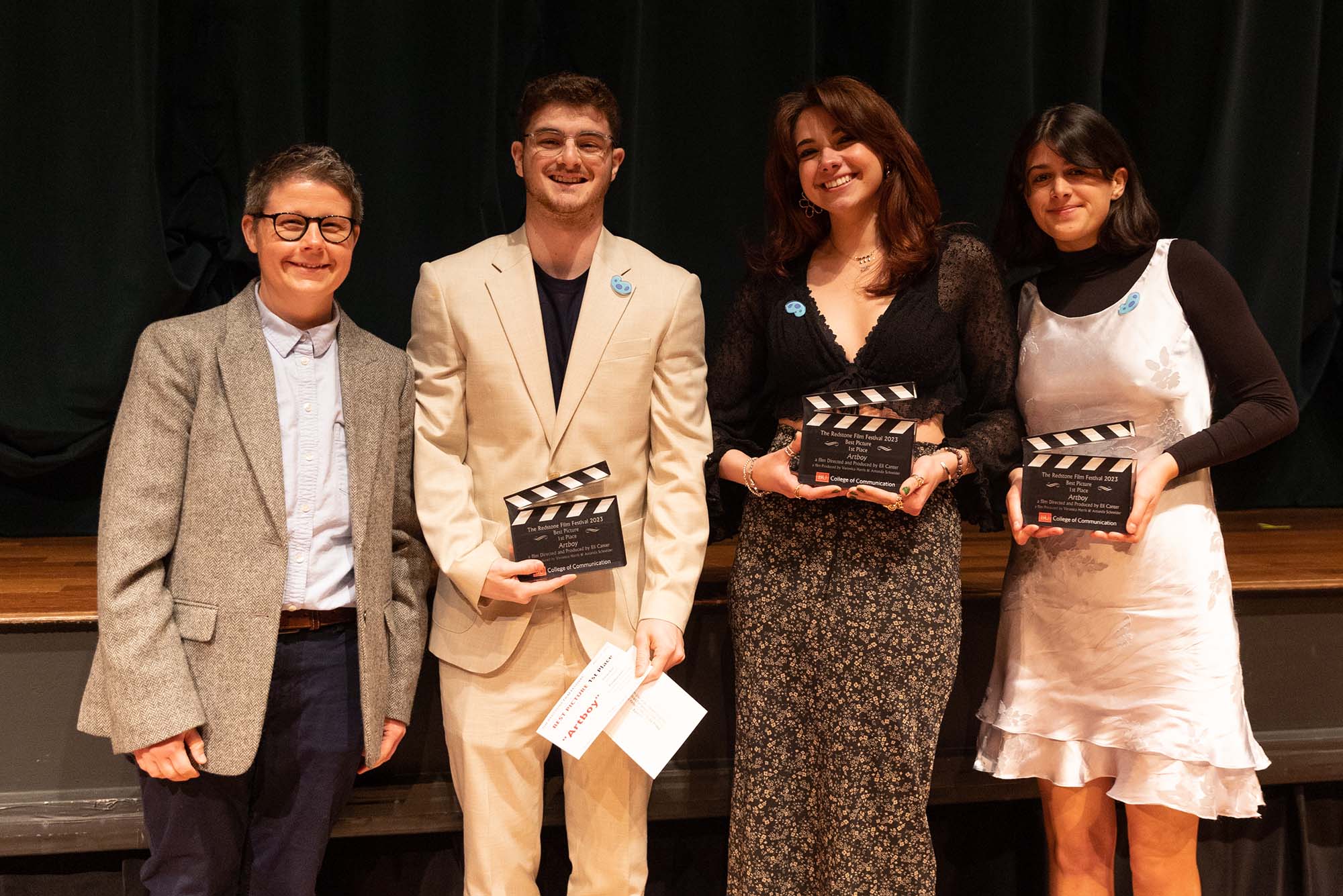 Photo: A group shot of the winners of the Redstone Film Festival. From left, a short white man in a gray blazer and a blue button-up dress shirt. Next, a taller Southern Asian man in a stream-lined tan suit and a black t-shirt. Next to him, a Southern Asian woman in a black, lace top and a floral maxi skirt. And lastly, the person on the right is a Sourthern Asian woman with a black mock-neck top and a silk, grey dress.