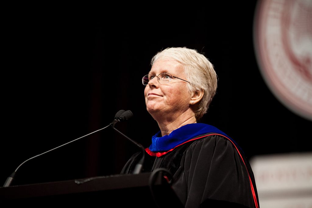 Photo: University Provost and Chief Academic Officer, Jean Morrison, an older white woman with short grey-brown hair, wears glasses and black ceremonial regalia. She has a blue collar and smiles as she looks up into the middle distance as she stands at a podium in front of a small microphone.