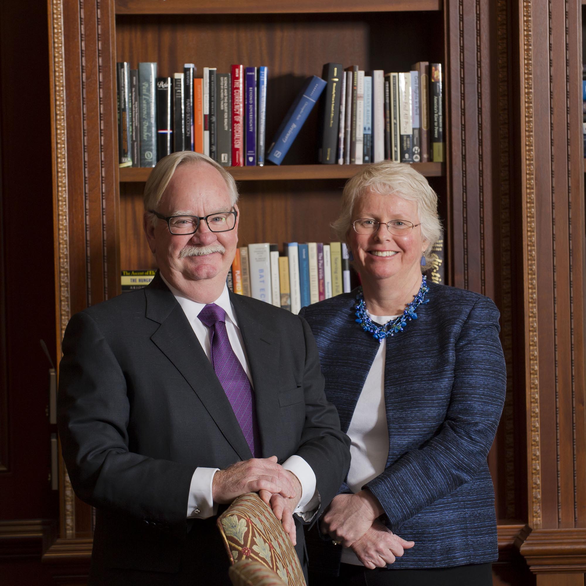 Photo: President Bob Brown and Provost Jean Morrison stand side by side and poses for a photo. A white man with receding grey hair and wearing glasses, a purple tie, and black suit stands to the left of a while woman with short grey hair wearing glasses, a white blouse, and blue tweed blazer. They both smile and stand with hands clasped in front of them and stand in front of a large wooden bookcase.