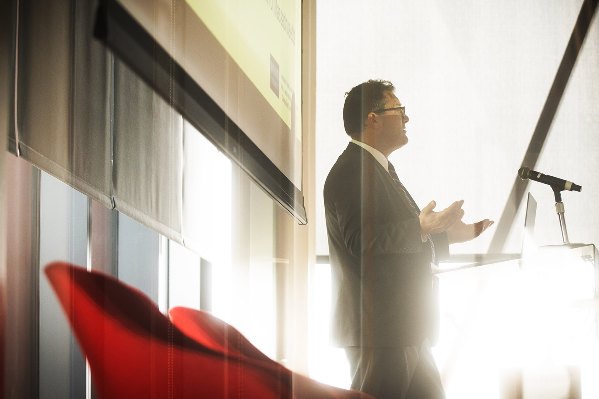 Photo: A man in a suit stands in front of a podium with a washed out white background and sun shining in through nearby windows