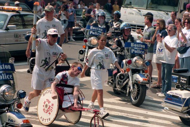 Photo: A race from 1996 featuring Rick and Dick Hoyt, father and son who competed in over 1000 races. They are in white shorts and a white t-shirt, the wheelchair that Rick is in has two big wheels on the side.