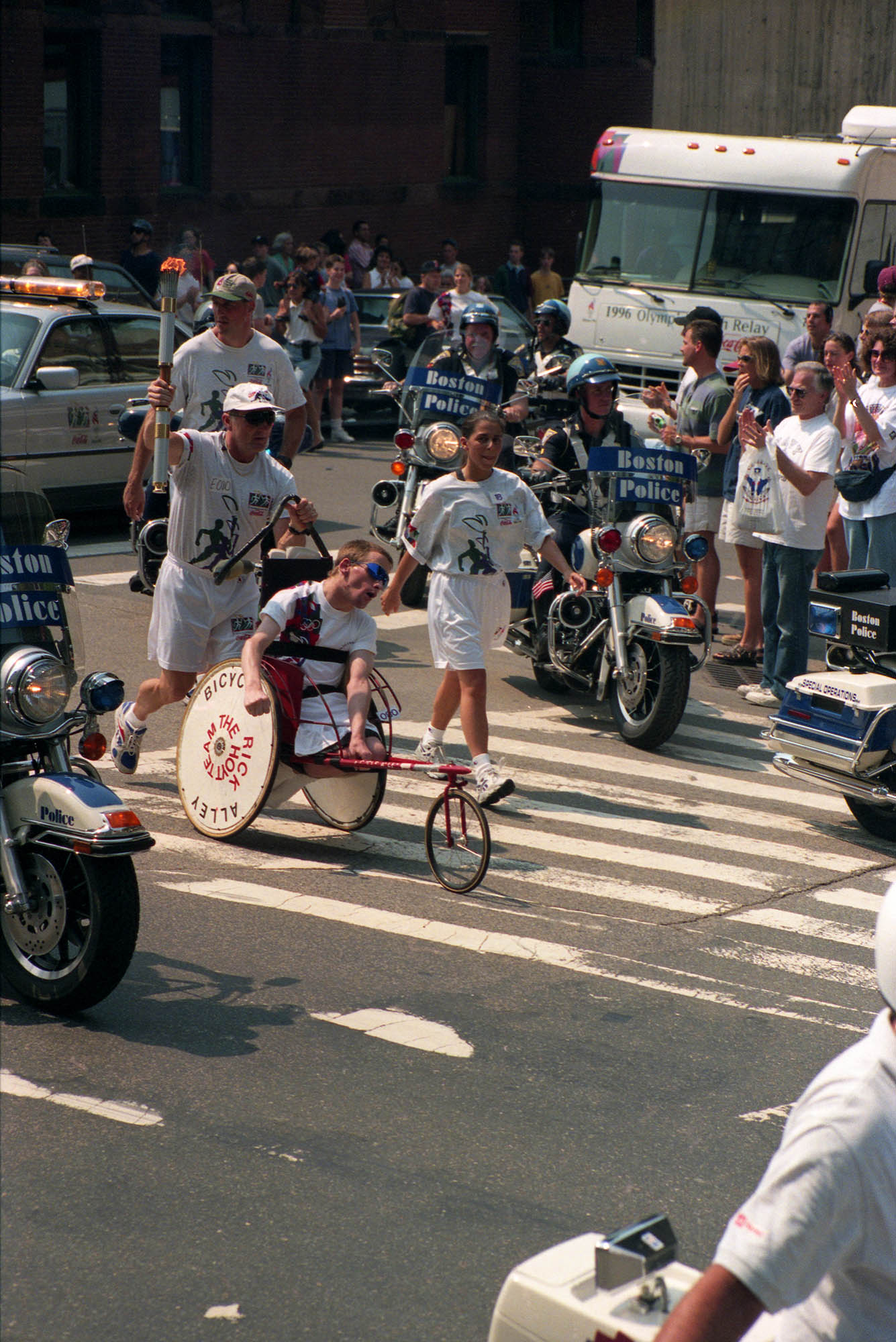 Photo: A race from 1996 featuring Rick and Dick Hoyt, father and son who competed in over 1000 races. They are in white shorts and a white t-shirt, the wheelchair that Rick is in has two big wheels on the side.