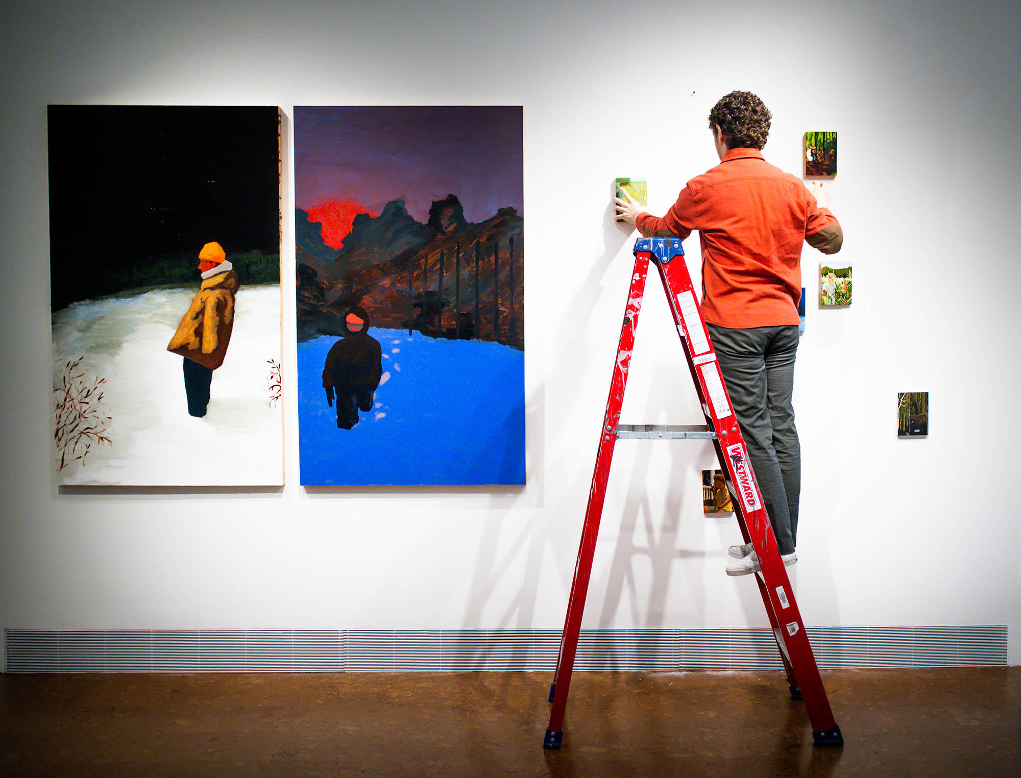 Photo: A person wearing an orange shirt and grey pants stands on a red ladder as they hang up large canvas paintings on a white wall.