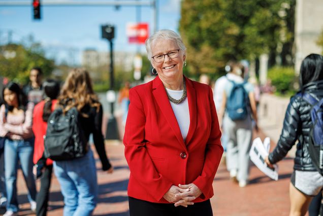 Photo: Portrait of Boston University Provost Jean Morrison on Commonwealth Ave. Asmiling white woman with short, silver white hair and wearing glasses, a white blouse, and red blazer poses with fingers clasped in front of her on a bustling sidewalk. Background and passerby are blurry.
