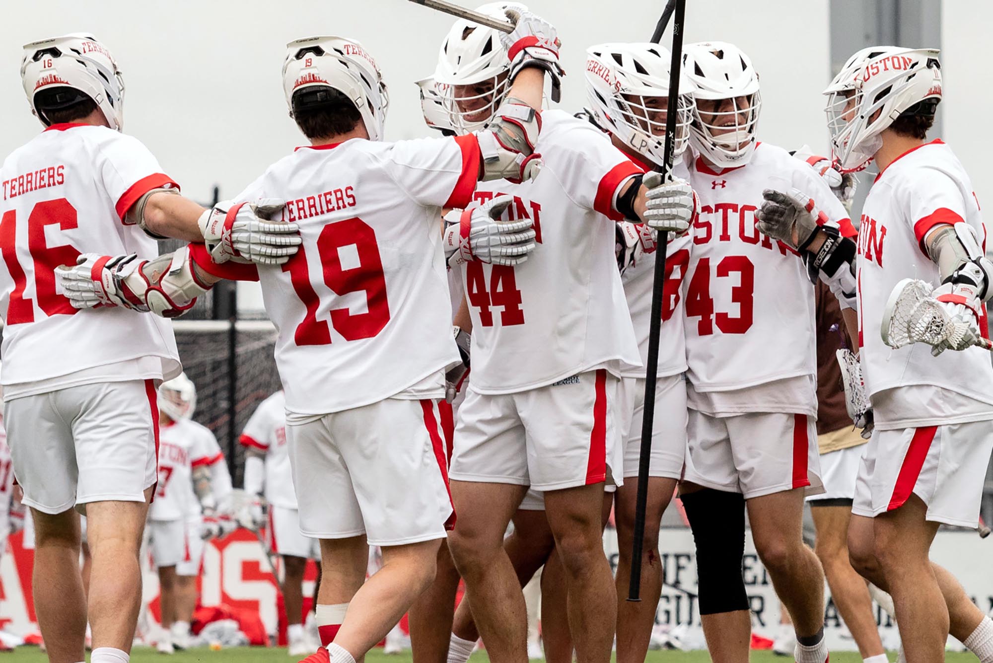 Photo: A group of BU lacrosse team members celebrate in a huddle. They are wearing their white uniforms with red detailing and are playing on in inside field.
