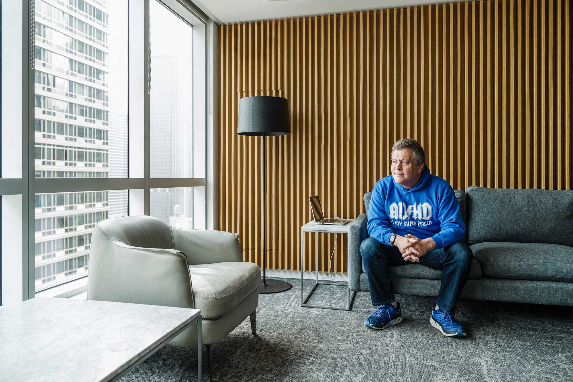 Photo: A man in a blue sweater sits on a modern leather couch staring out the window in NYC. Peter Shankman (CGS’92, COM’94), author of a new children's book on neurodiversity. Photo by Chris Sorensen for Boston University Photography.