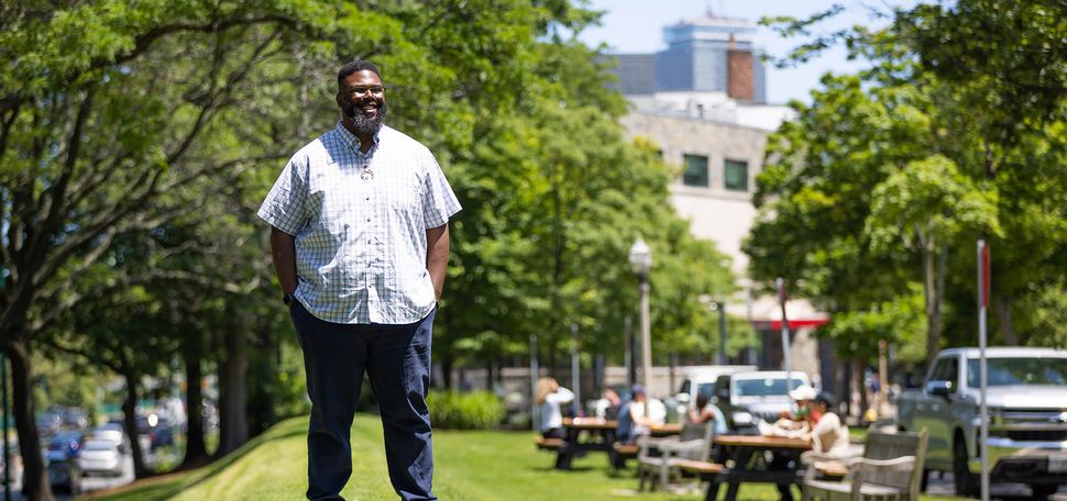 Photo: Anthony (Tony) Jack, a Black man wearing glasses, a light plaid collared shirt, and jeans, stands on a small grassy hill on a sunny day. Picnic tables filled with people can been in the blurry background behind him.