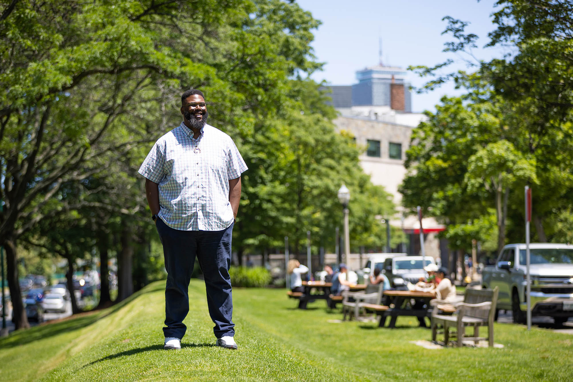 Photo: Anthony (Tony) Jack, a Black man wearing glasses, a light plaid collared shirt, and jeans, stands on a small grassy hill on a sunny day. Picnic tables filled with people can been in the blurry background behind him.