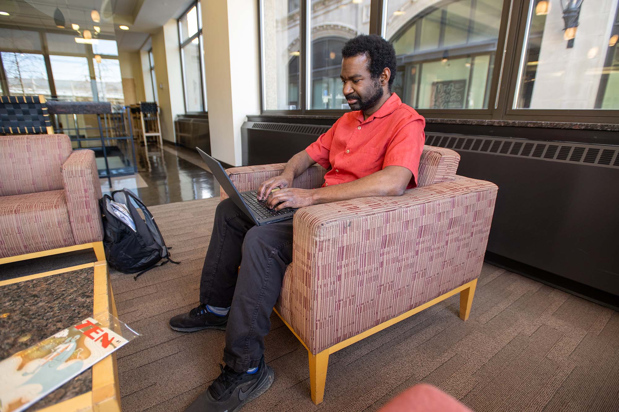 Photo: Chris Rhodes, a Black man wearing a red short-sleeved collared shirt and black pants, sits in an armchair as he works.