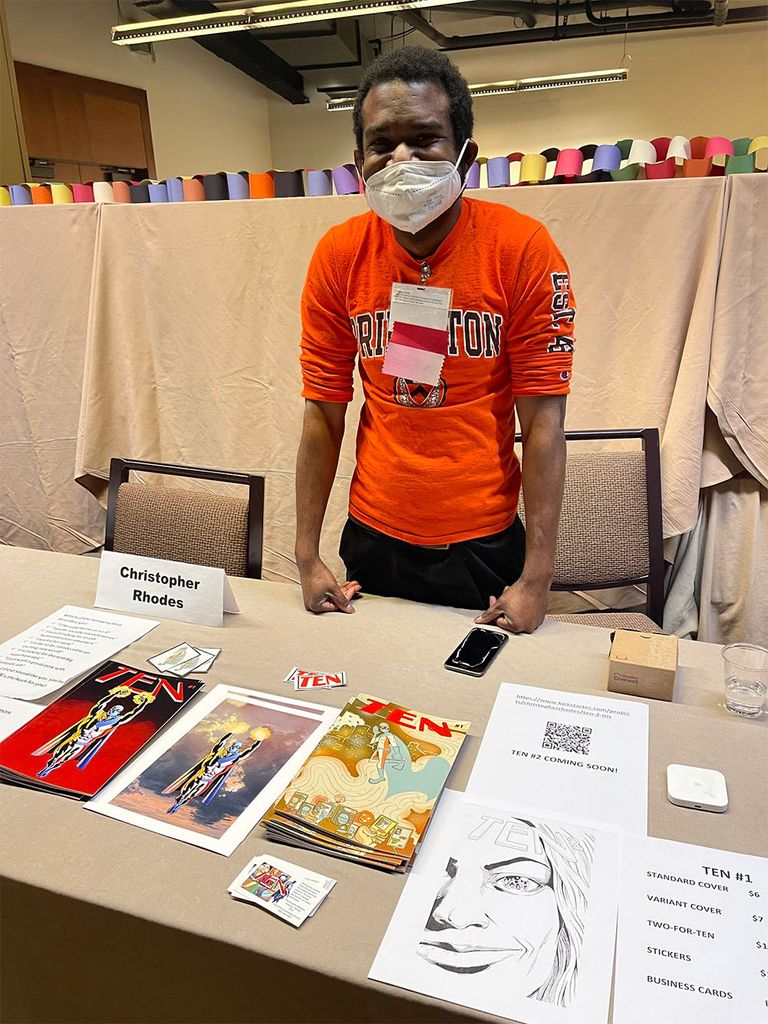 Photo: Rhodes, a Black man wearing a red shirt, face mask, and jeans stands behind a table with three comic books on display.