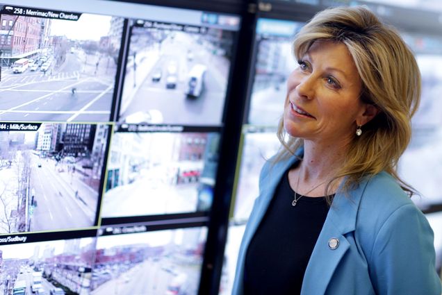 Photo: Gina Fiandaca, a white woman wearing a black shirt and light blue blazer, is shown looking at various screens of video footage of various MBTA stations.