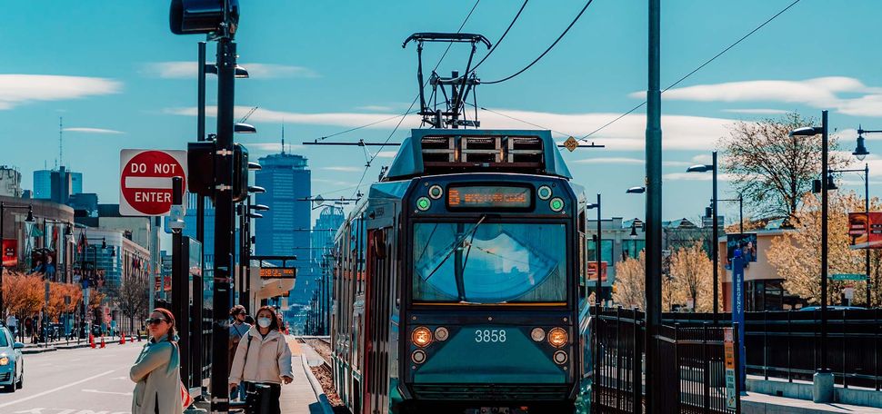 Photo: The MBTA Green B Line Trolley is shown on commonwealth Avenue. People stand on the sidewalk to the left as the T passes to the right. The Boston University and Boston city skyline can be seen i the distance on the sunny day.