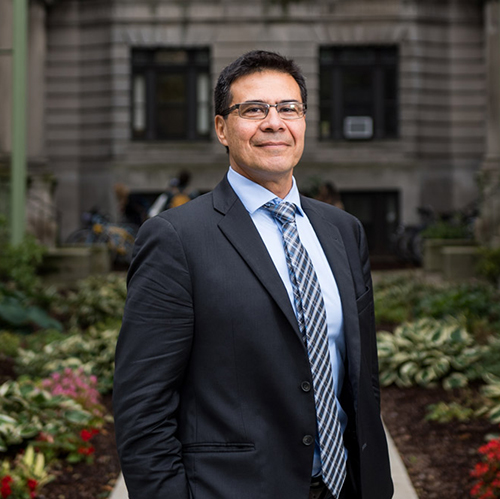 Photo: Jorge Delva, a tan man wearing glasses, a light blue collared shirt, black blazer, and striped blue tie, smiles and poses outside. A grey brownstone and flowers can be seen behind him in ht blurry background.