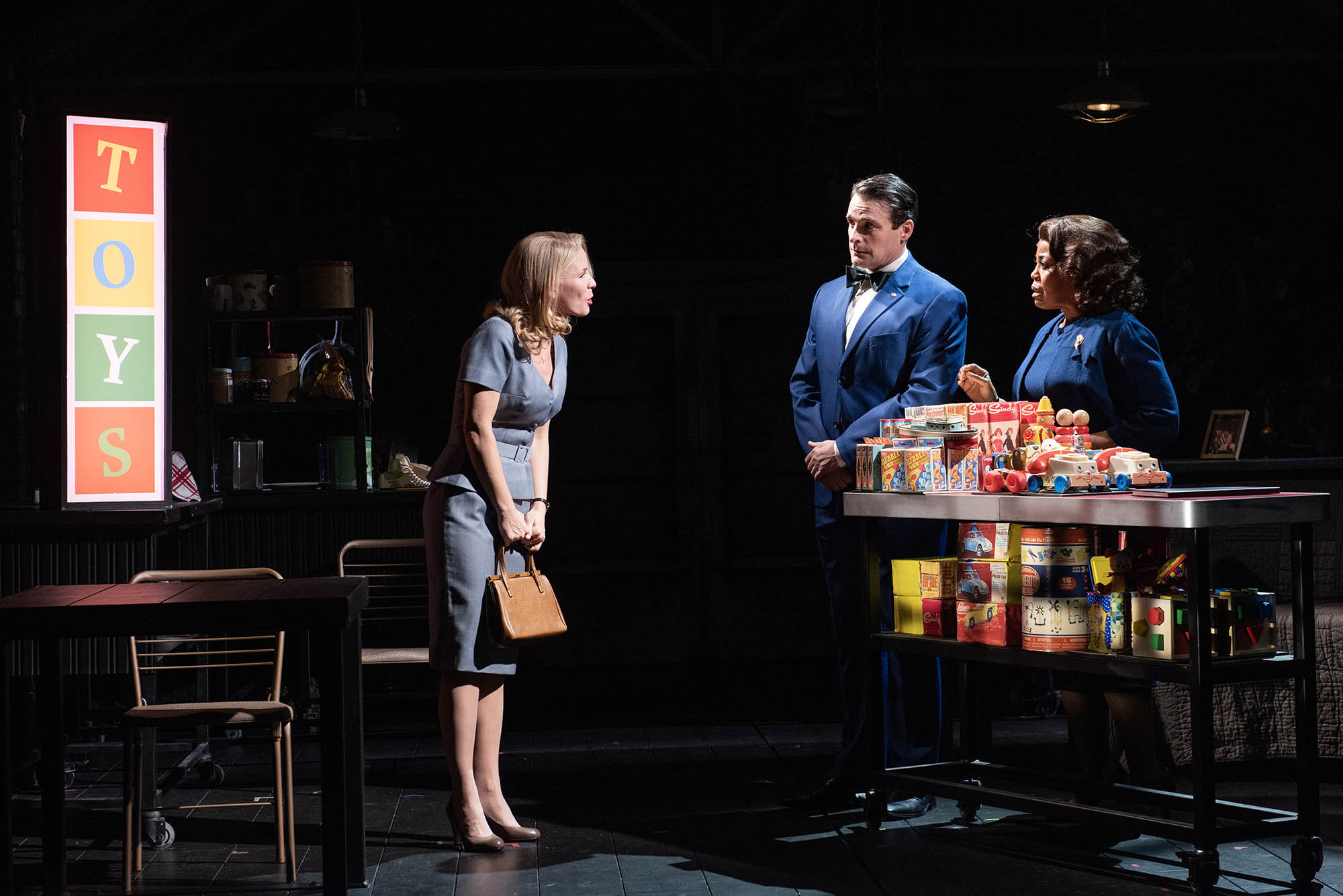 Photo: Kelli O’Hara as Kirsten is buying a toy. A white woman wearing a light blue dress talks to two store clerks standing in front of a table full of toys to her right on stage. A spotlight highlights the scene on the otherwise dark stage.
