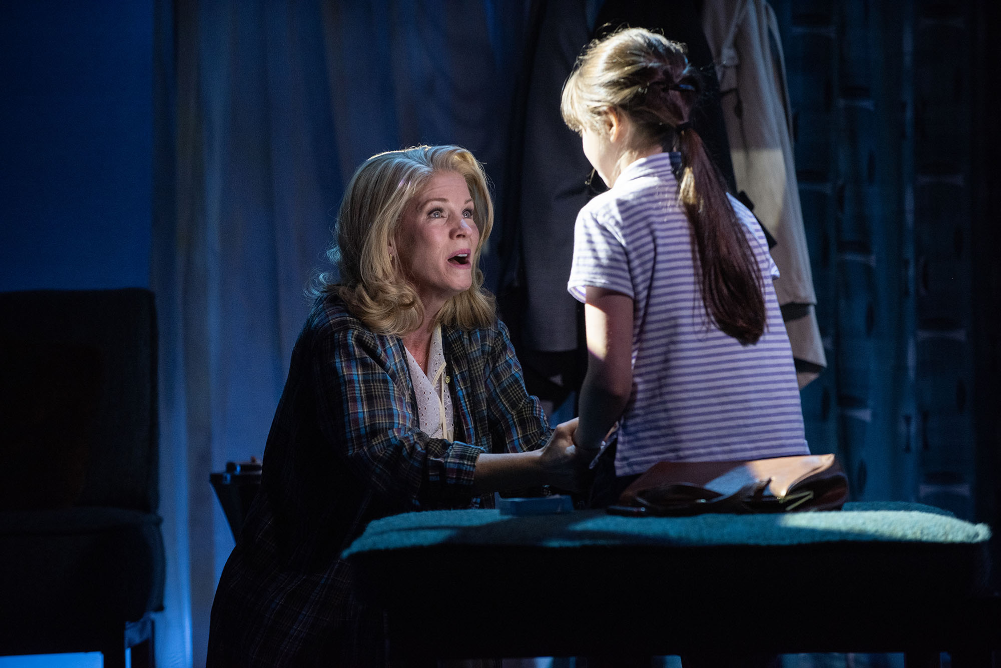 Photo: O’Hara with Ella Dane Morgan as daughter Lila. A white woman dressed in pajamas bends down and sings to a young white child. A spotlight illuminates them on an otherwise dark stage.