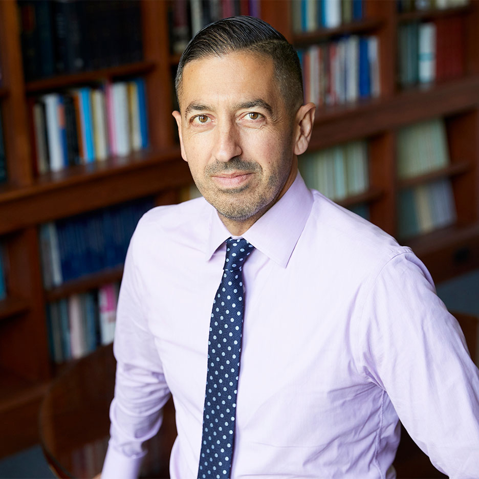 Photo: headshot of Dean Sandro Galea, a tan man wearing a lavender, long-sleeved collared shirt and blue and white polka dot tie. Wooden bookshelves filled with books are seen in the blurred background.